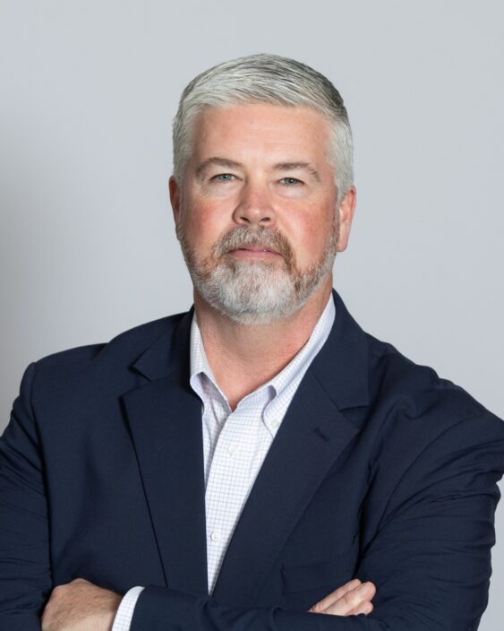 Professional portrait of a middle-aged man with gray hair and beard, wearing a navy blazer and light checkered shirt, arms crossed against a neutral gray background